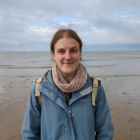 Corinne is standing on the beach with the sea in the background. She is wearing a blue jacket and pink scarf.
